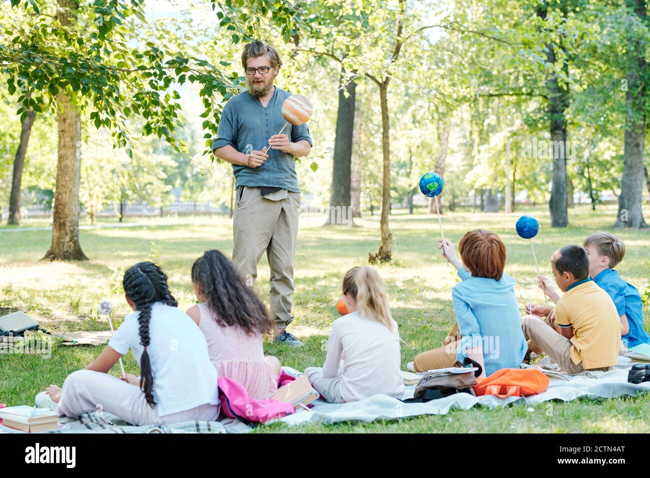 Maestro barbudo sosteniendo modelo de planeta en palo y hablando con grupo de niños sentados en el suelo del parque Foto de stock