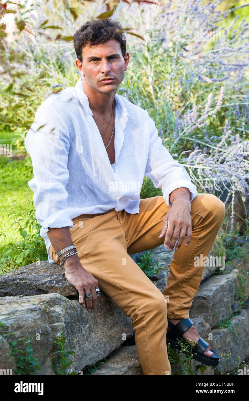 Longitud completa de hombre joven feliz en camisa blanca sin botones y  coloridos jeans con accesorios sentados en el borde de piedra jardín verde  de verano Fotografía de stock - Alamy