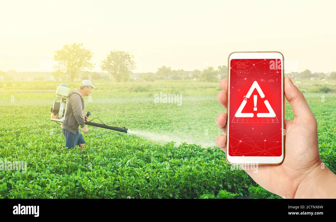 Teléfono en la mano mostrando una advertencia contra el fondo de un agricultor con un nebulizador nebulizador nebulizador en un campo agrícola. Uso de productos químicos peligrosos para el tratamiento Foto de stock