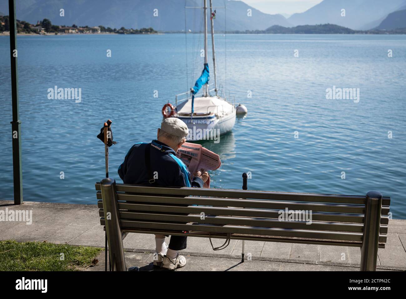 El anciano se relaja en un banco mientras lee el periódico en el lado del lago como en Gravedona, Lombardía, Italia. Foto de stock