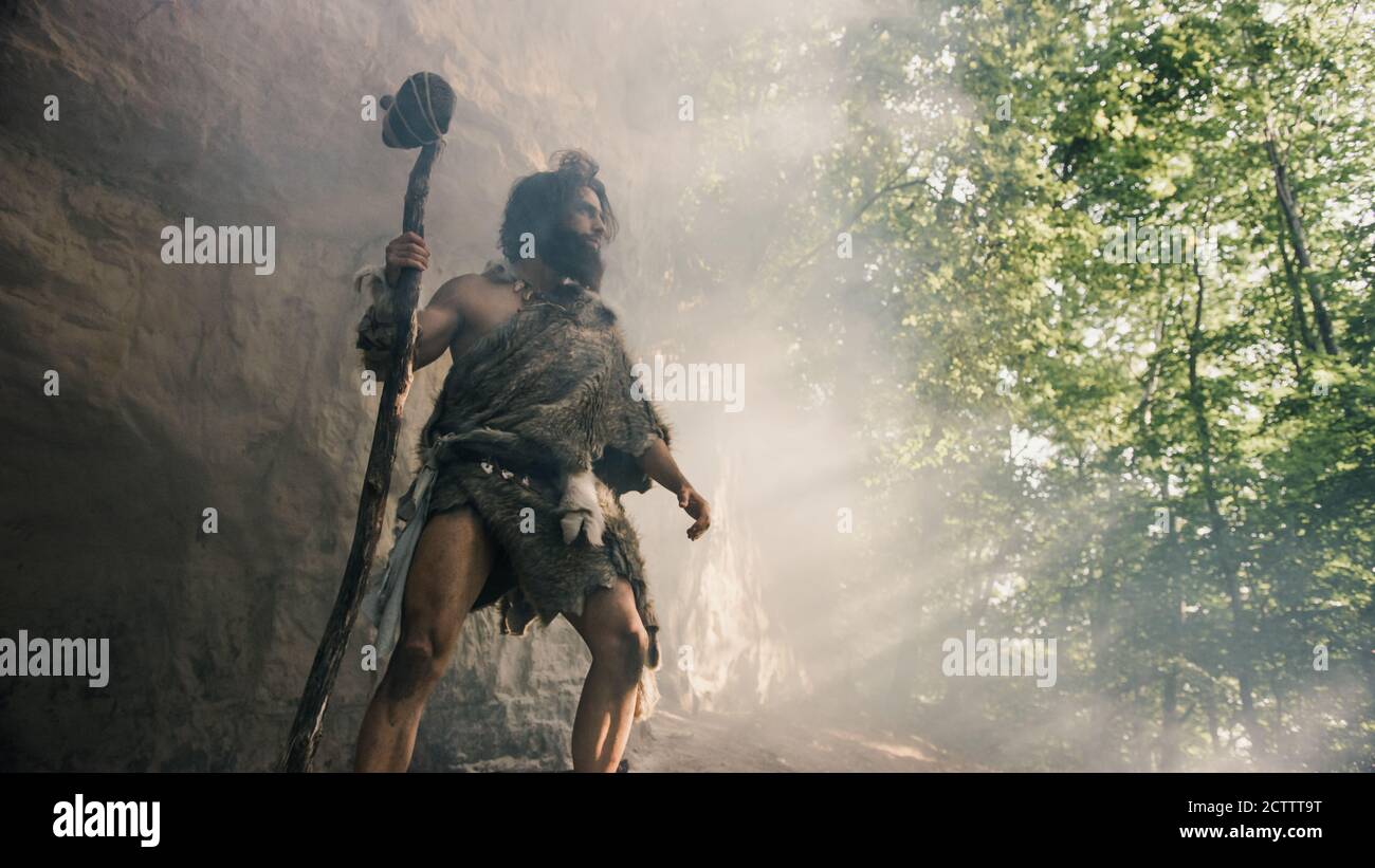 Primigenia Caveman que lleva piel animal sostiene martillo de piedra se encuentra cerca de la cueva y mira alrededor del paisaje prehistórico, listo para cazar animales de presa Foto de stock