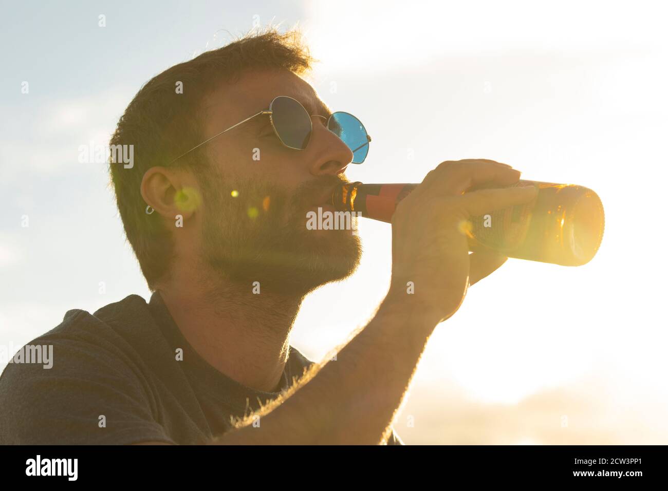 Un joven, con vasos y barba, bebe cerveza de una botella, mientras disfruta del paisaje y la puesta de sol, en Galicia, al norte de España. Foto de stock