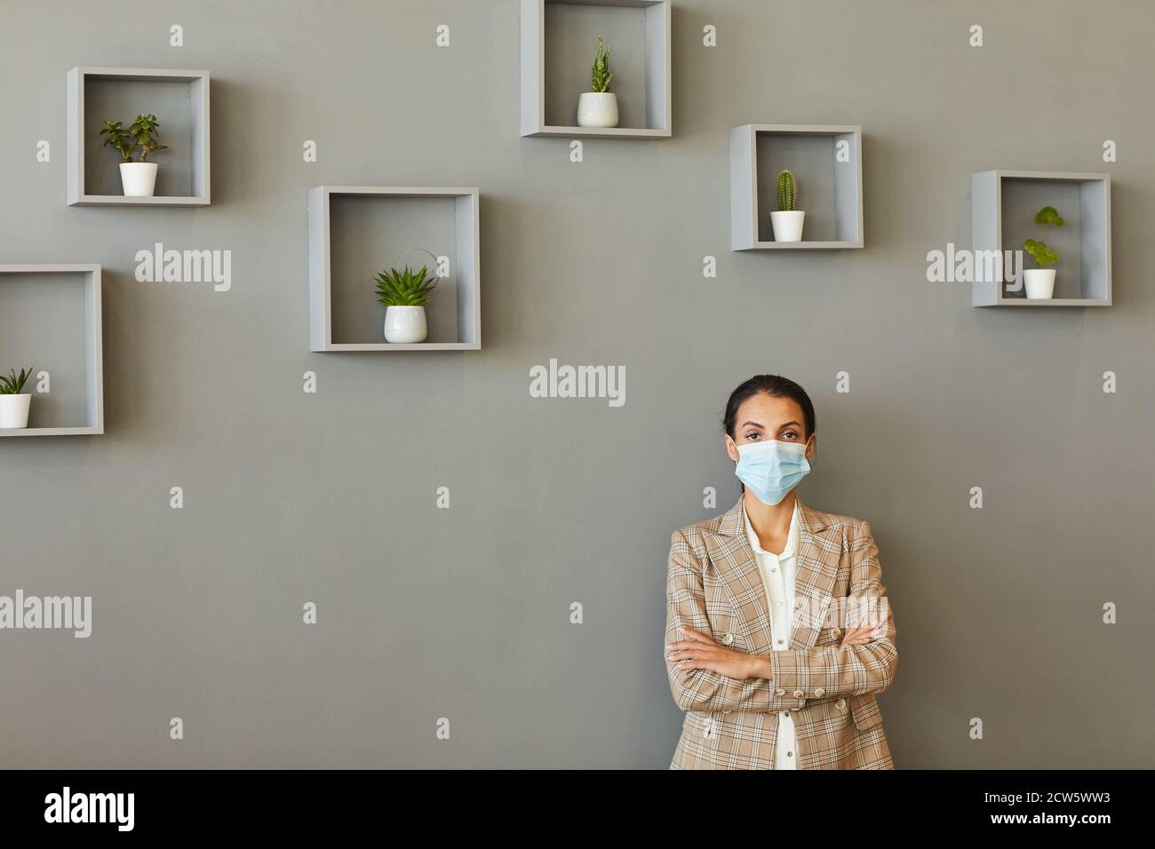 Retrato mínimo de una mujer de negocios segura con máscara mientras está de pie con los brazos cruzados contra la pared gris, espacio de copia Foto de stock