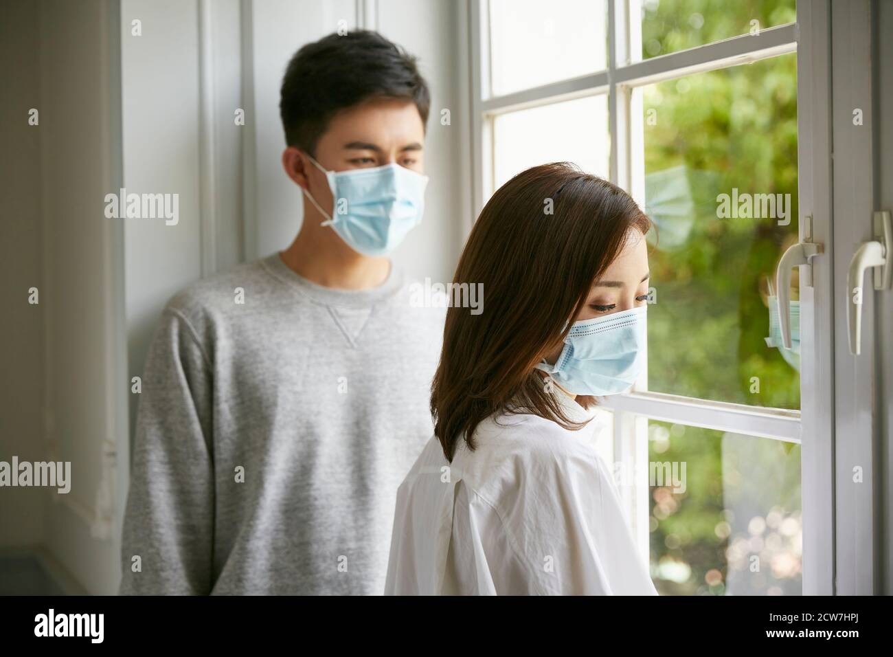 joven pareja asiática en aislamiento con máscara de pie junto a la ventana Foto de stock