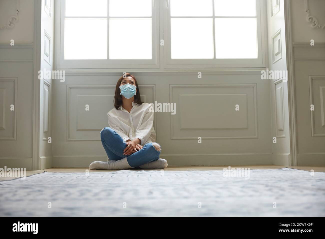 joven asiática en cuarentena en casa sentada en el suelo piernas vestida con mascarilla facial Foto de stock