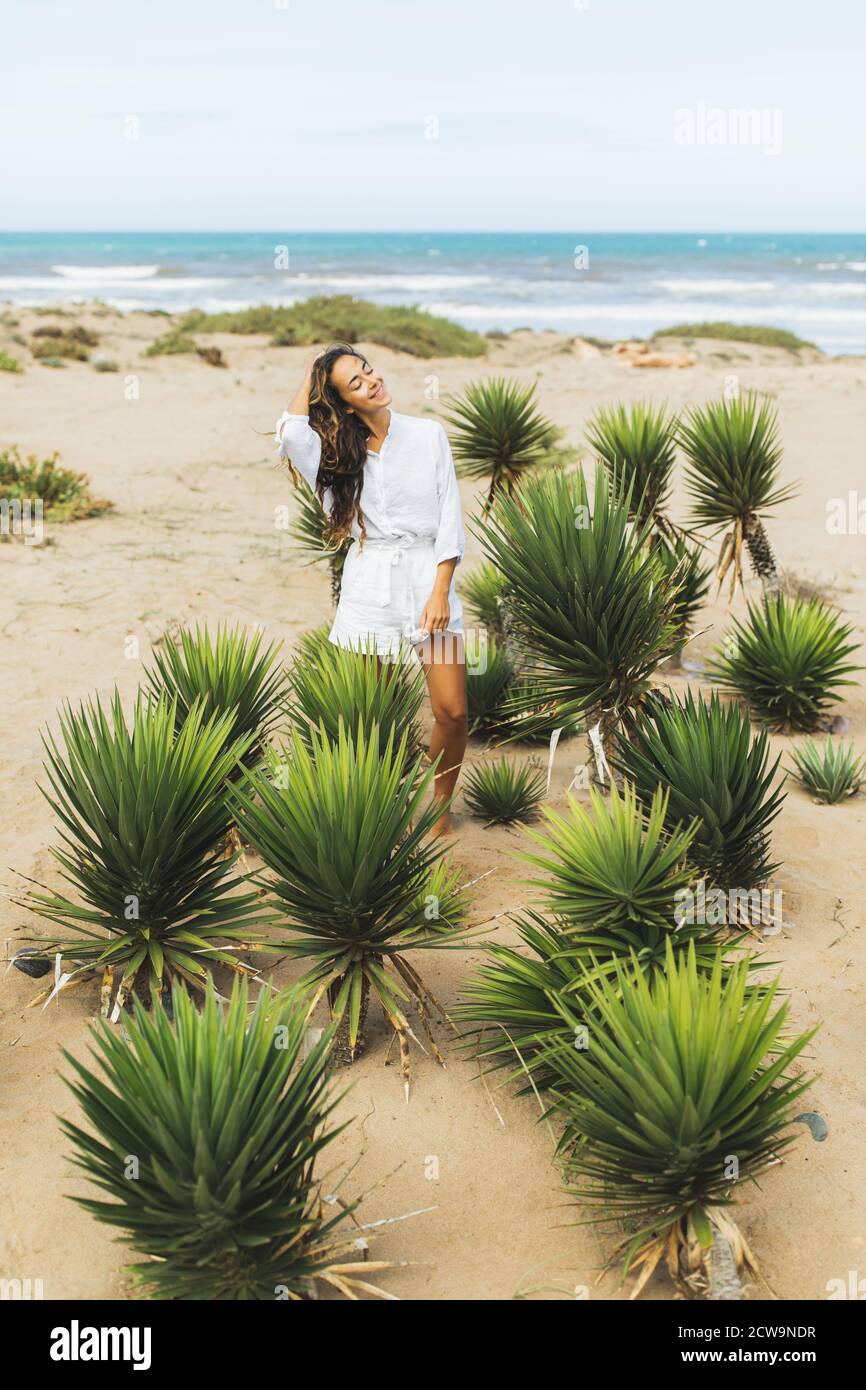 Mujer feliz sonriendo con camisa blanca y pantalones cortos en la playa de arena del océano. Plantas de cactus verdes tropicales. Tiempo de viento. Foto de stock