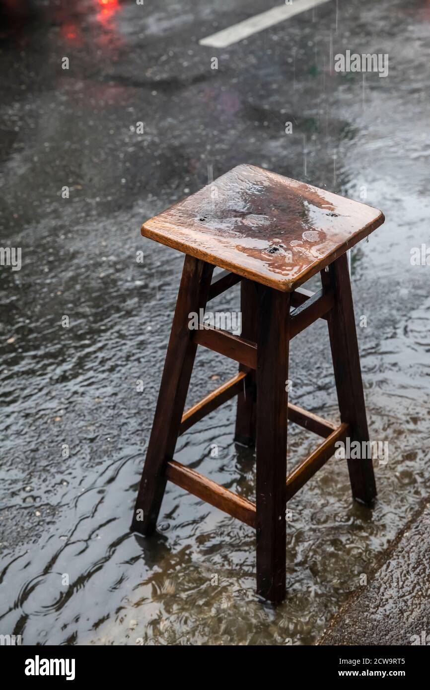 Un taburete de madera se sienta bajo la lluvia en el borde de una calle concurrida sin ningún propietario a la vista en el barrio chino de Bangkok. Foto de stock