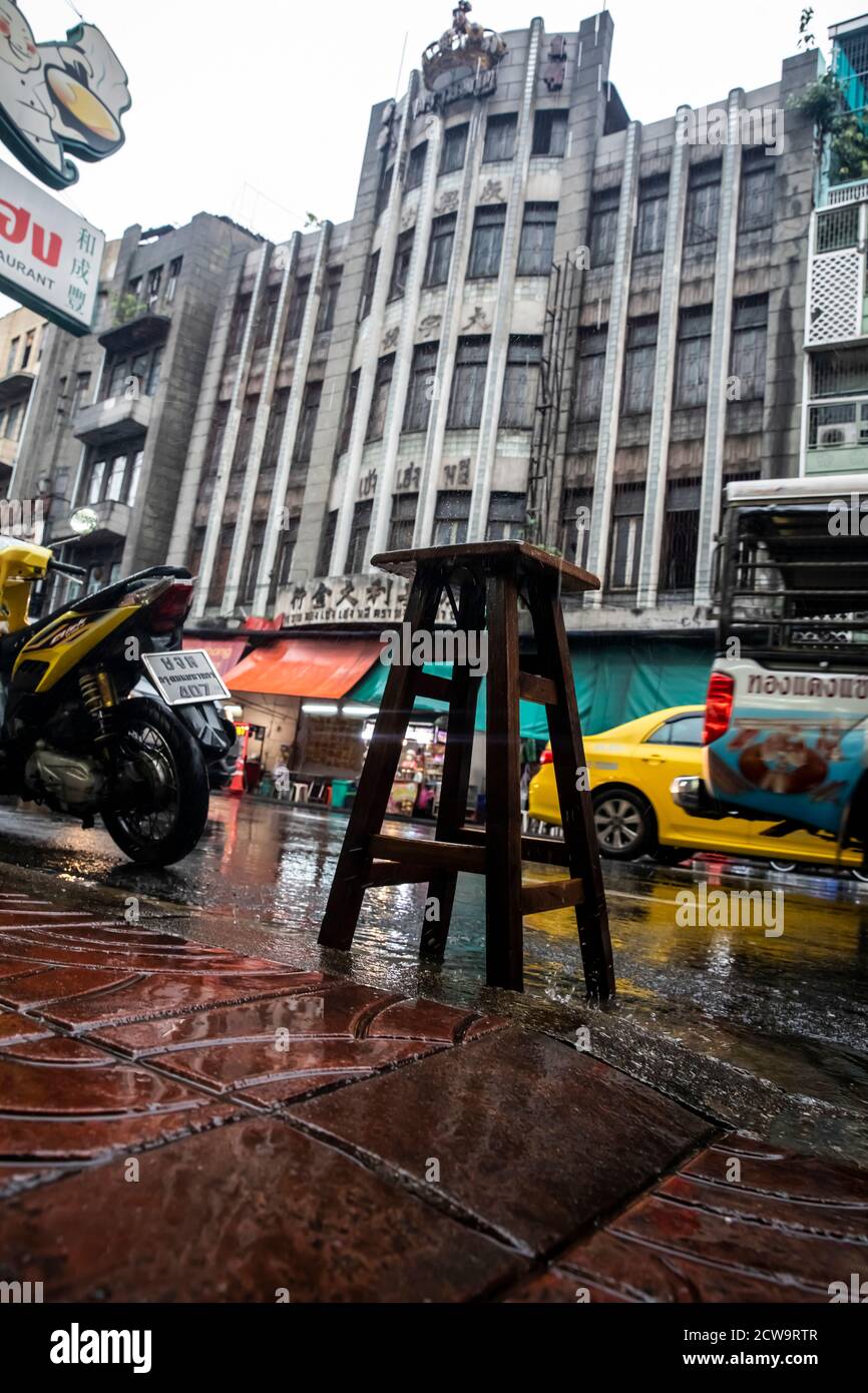 Un taburete de madera se sienta bajo la lluvia en el borde de una calle concurrida sin ningún propietario a la vista en el barrio chino de Bangkok. Foto de stock