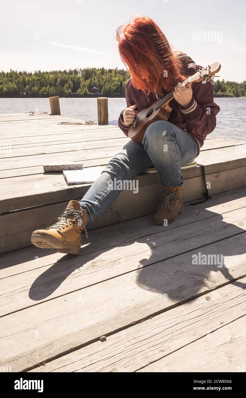Una niña adolescente de pelo rojo canta una canción con ukulele sentado en un muelle de madera en un día soleado Foto de stock