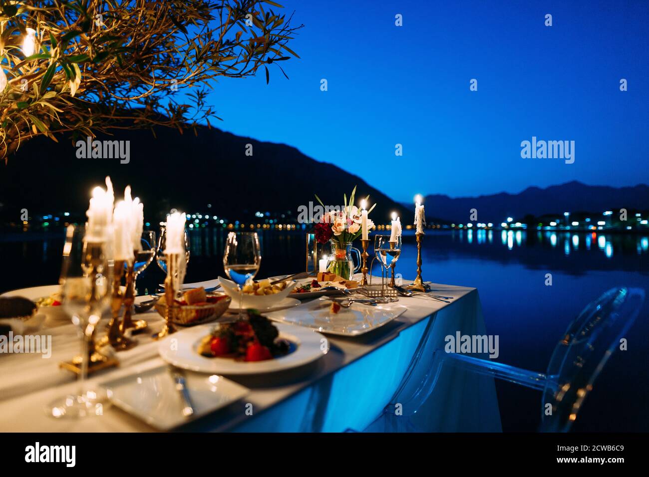 Una mesa con cena para dos, un ramo de flores y velas en el fondo de agua y  montañas por la noche, en un restaurante de la ciudad Fotografía de stock -