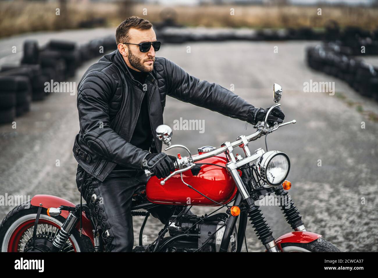 Moto roja con jinete. Un hombre en una chaqueta de cuero negro y pantalones  está de lado en el medio de la carretera. Los neumáticos se colocan sobre  el fondo Fotografía de