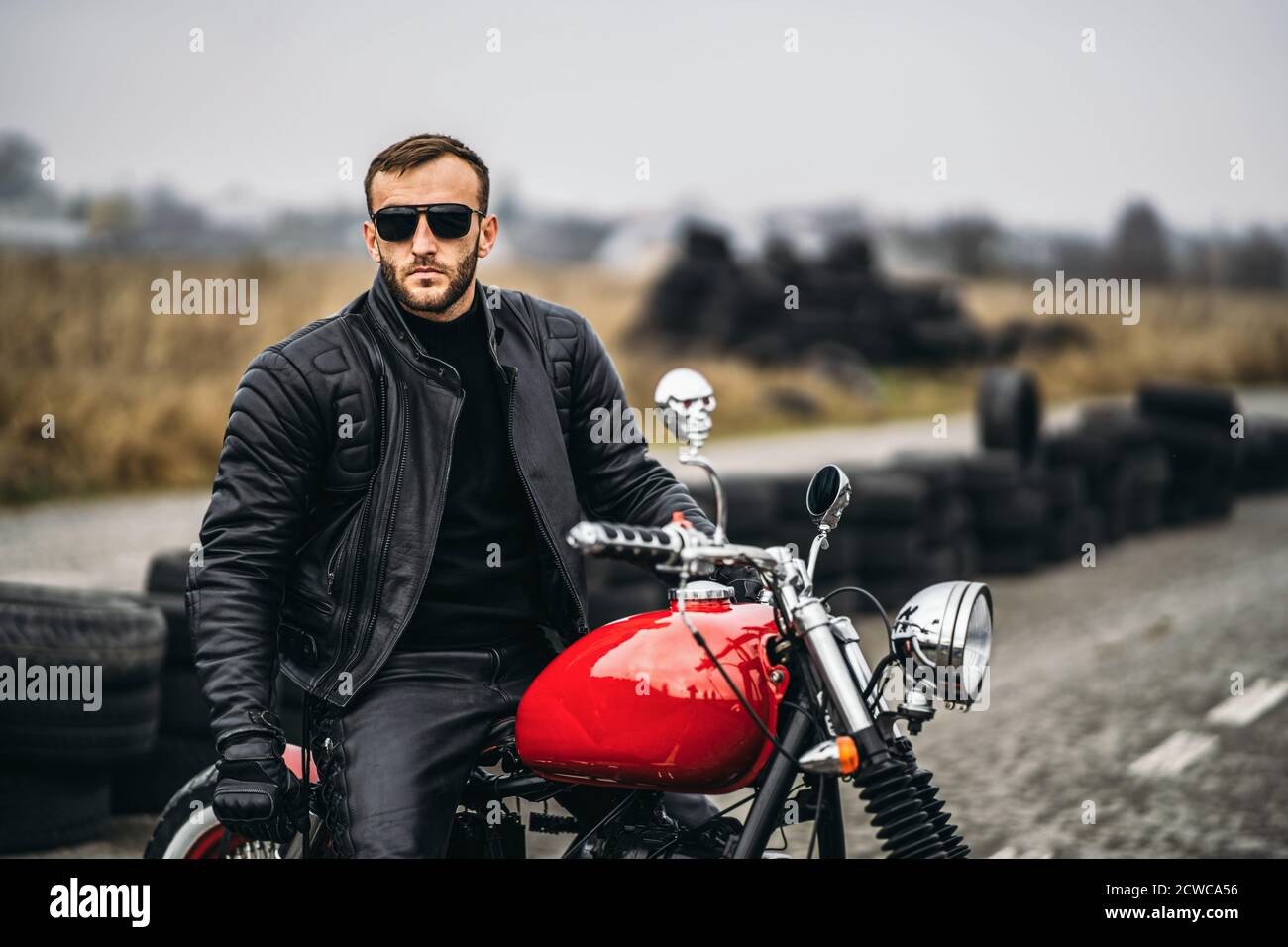 Moto roja con jinete. Un hombre en una chaqueta de cuero negro y pantalones  está de lado en el medio de la carretera. Los neumáticos se colocan sobre  el fondo Fotografía de