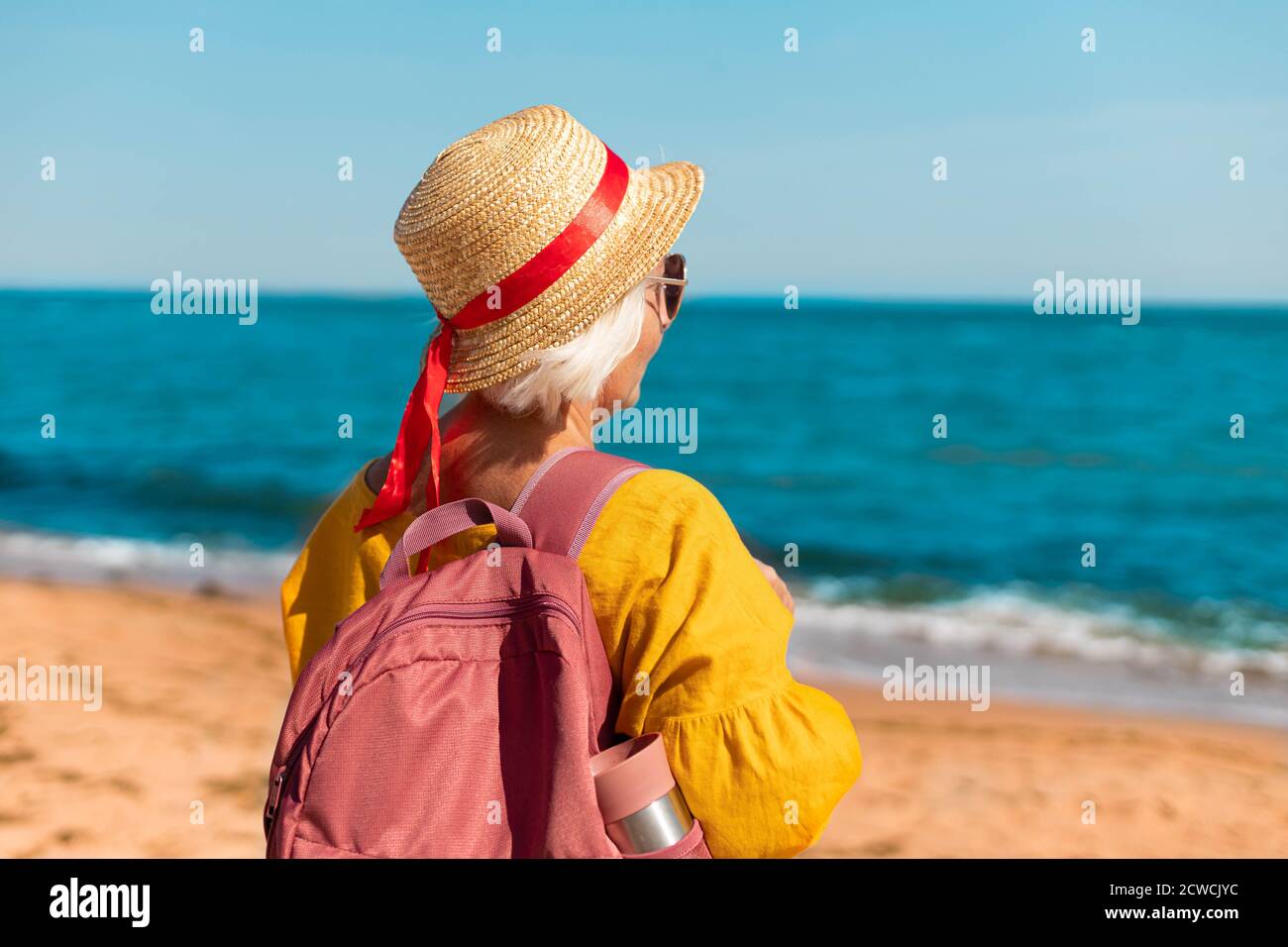 Retrato de una joven feliz en un pajizo de verano sombrero y gafas con mochila turística y termo disfrute de la hermosa vista al mar Foto de stock