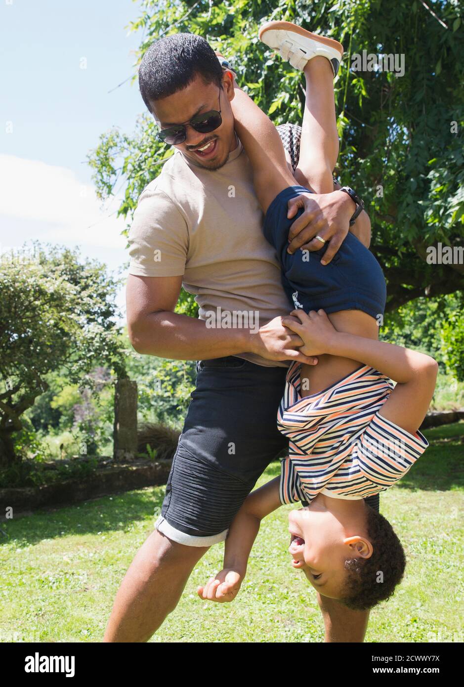 Padre juguetón sosteniendo al hijo al revés en el soleado patio trasero de verano Foto de stock