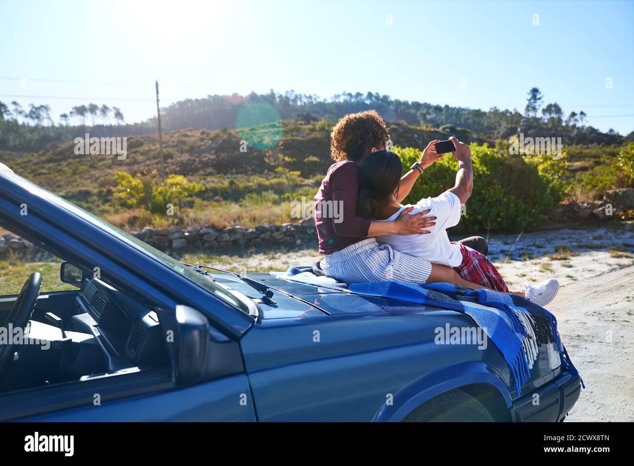 Una pareja joven despreocupada tomando selfie en la capucha del coche en carretera soleada Foto de stock