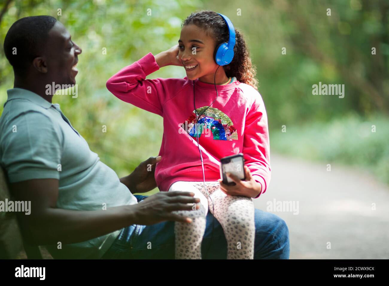 Feliz padre e hija sentados en el banco del parque con auriculares Foto de stock
