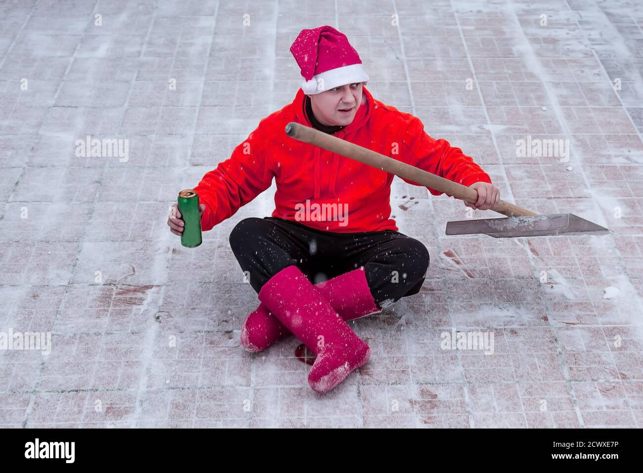 Hombre en un sombrero rosa de Navidad y botas de invierno en un suéter rojo  está sentado en una posición de loto en el sitio de su patio con una pala.  Él