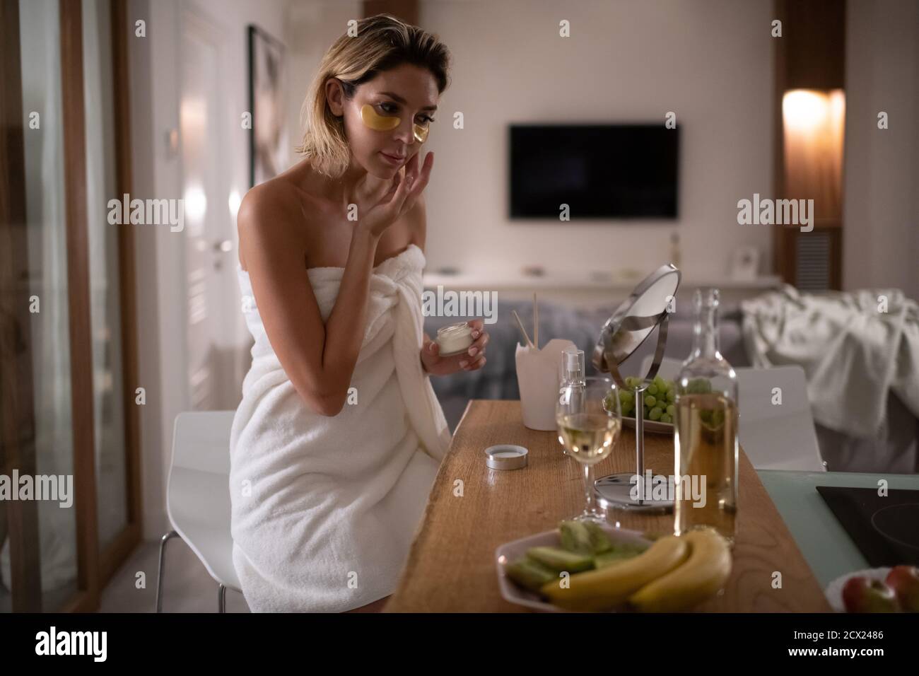 Mujer adulta aplicando crema en la mejilla mientras se sienta en la mesa después de cenar en casa Foto de stock