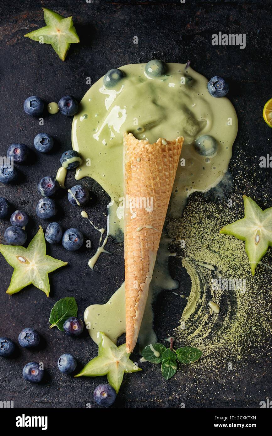 Cono de waffle de fusión del té verde matcha elástico suave helado con hojas de menta, carambolas, cal, arándanos negros sobre fondo de textura. Vista desde arriba. Foto de stock