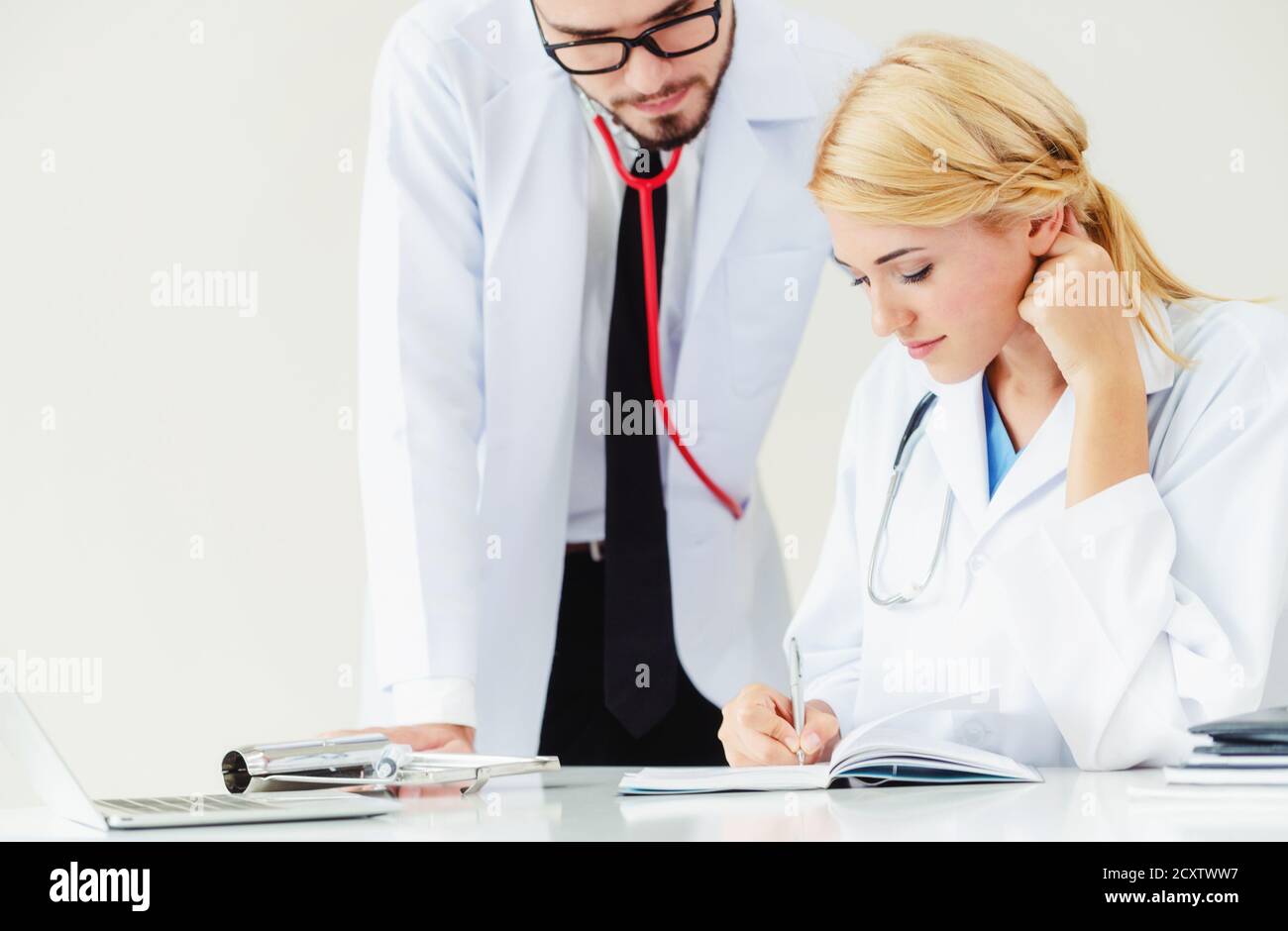 Doctor en el hospital office escribe notas sobre los pacientes informan al tener una conversación con otro médico que de pie junto a ella. Foto de stock