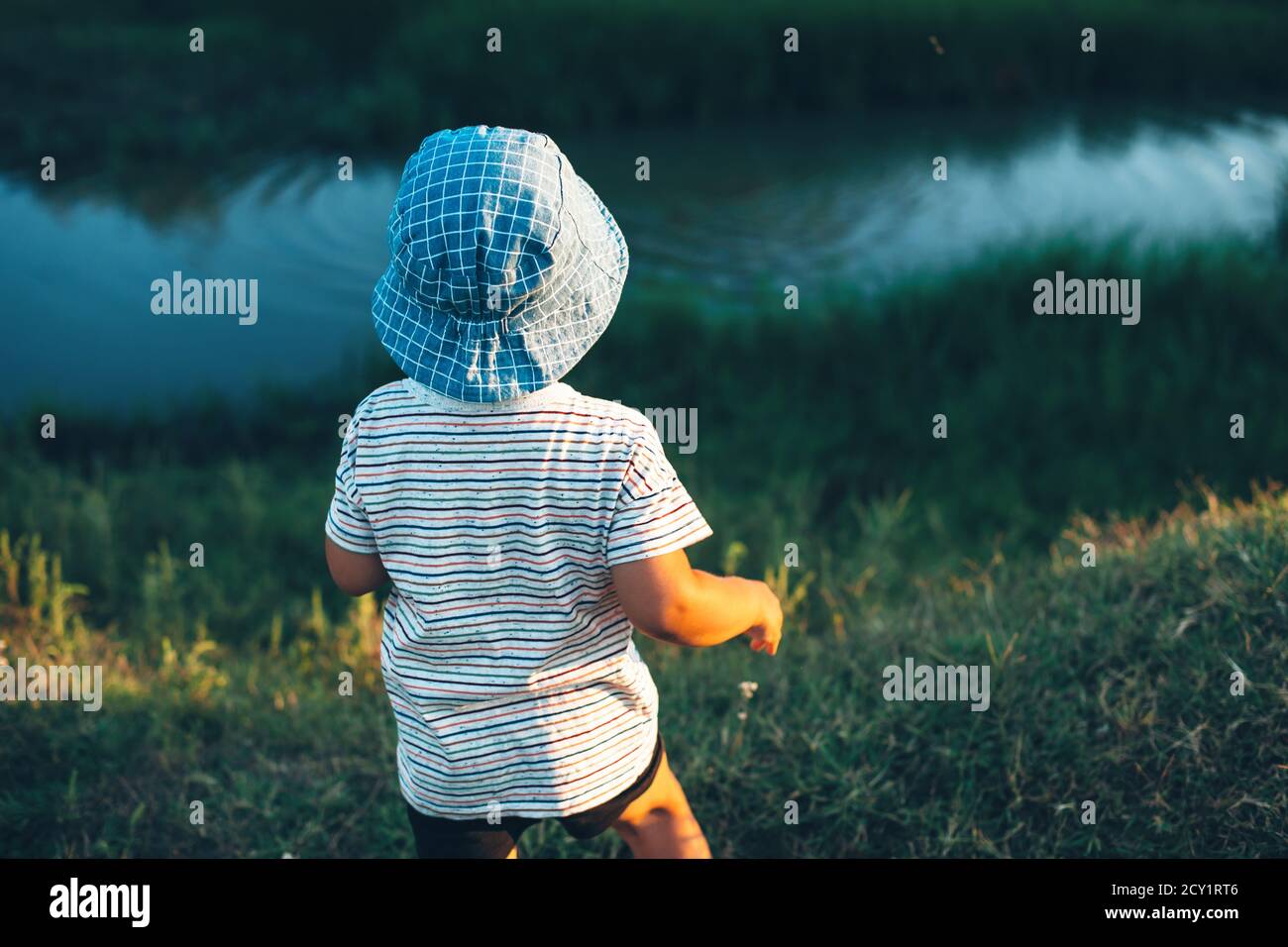 Niño caucásico mirando un pequeño río y lanzando piedras usando un sombrero azul en un campo verde Foto de stock