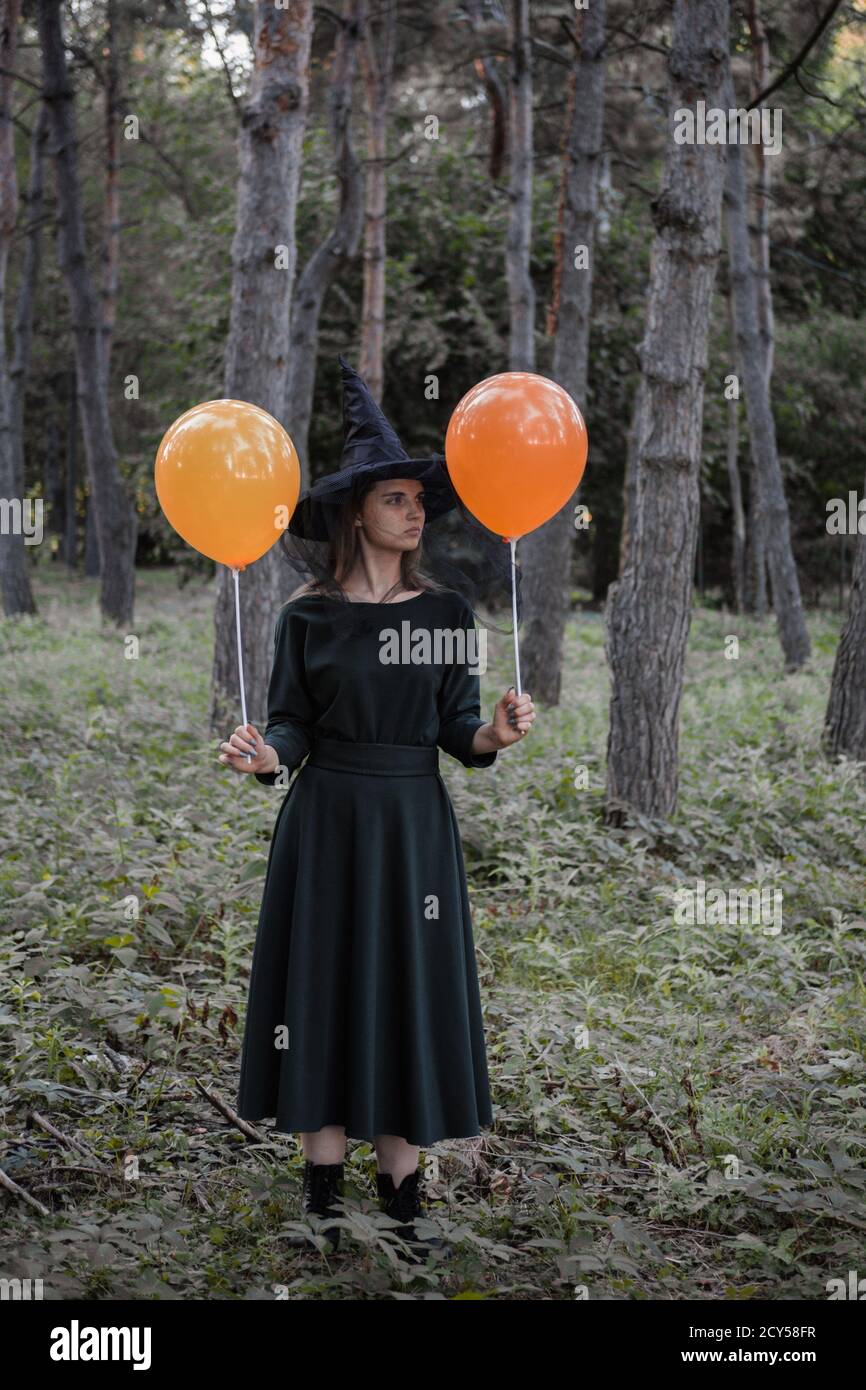 Joven linda hermosa mujer en vestido oscuro y sombrero de bruja sostiene  globos de naranja en sus manos. Disfraz de fiesta de Halloween. Bosque,  parque con árboles de otoño Fotografía de stock -