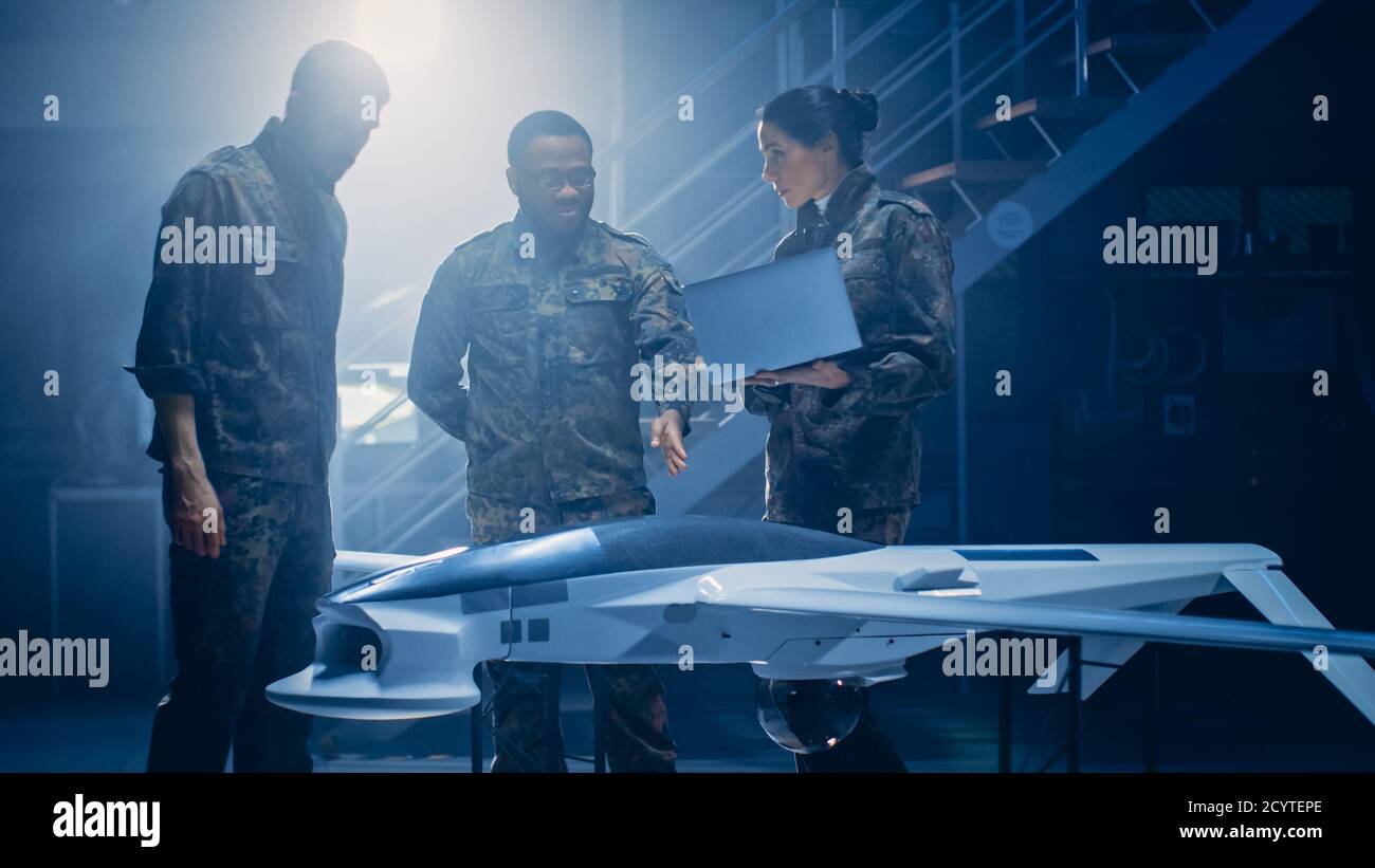 Los Ingenieros Aeroespaciales del Ejército trabajan en el vehículo aéreo no tripulado Drone. Expertos en aviación uniformados hablan, usando un ordenador portátil. Instalación industrial con aviones para Foto de stock