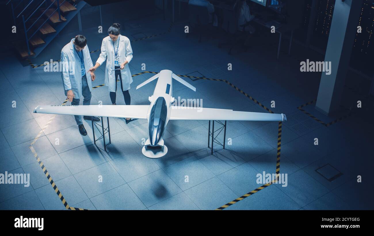 Dos ingenieros aeroespaciales trabajan en el prototipo de vehículo aéreo no tripulado Drone. Científicos de aviación en White Coats hablando, utilizando Tablet PC. Industrial Foto de stock