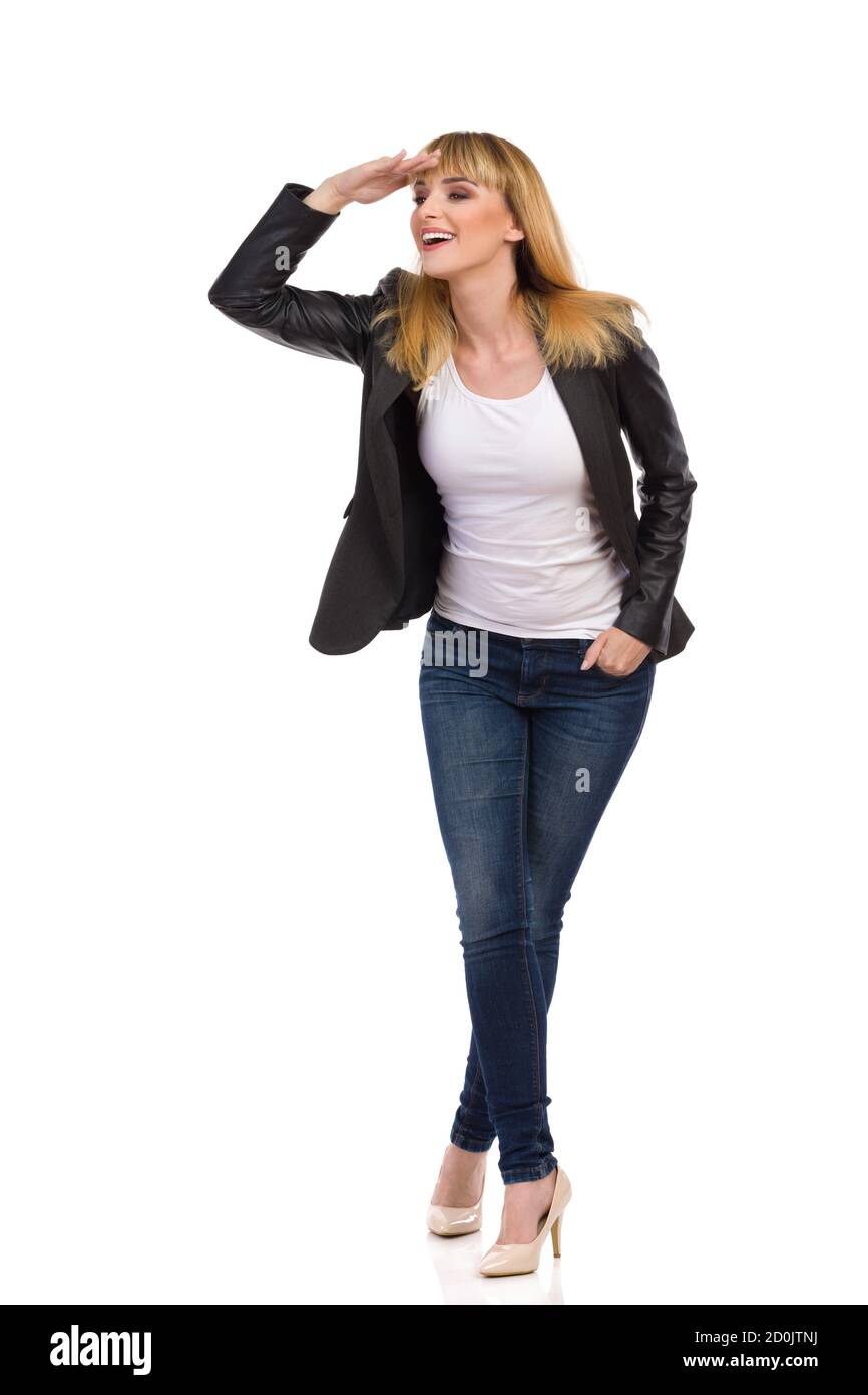 Joven mujer feliz en chaqueta de cola negra sin botones, jeans azules y tacones  altos está de pie, sosteniendo la mano en la frente y mirando hacia fuera.  Vista frontal. Lleno Fotografía