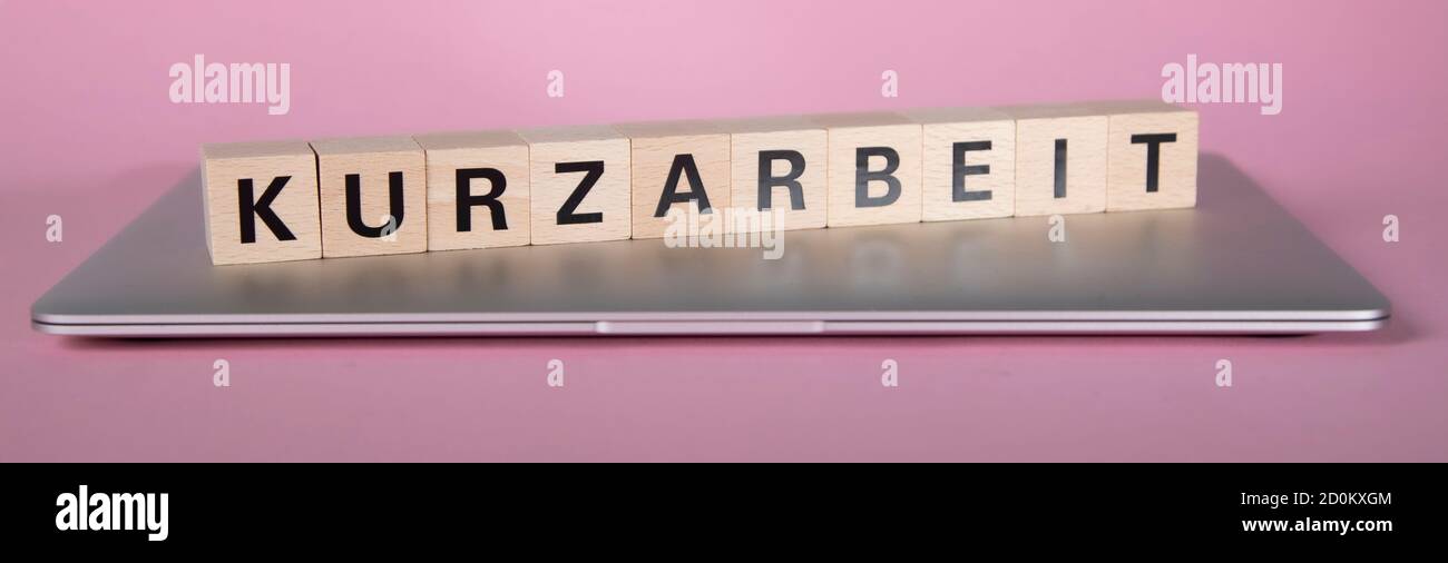Bloques de madera con palabra alemana para el trabajo de corto tiempo, Kurzarbeit, sobre un ordenador portátil cerrado, fondo rosa Foto de stock