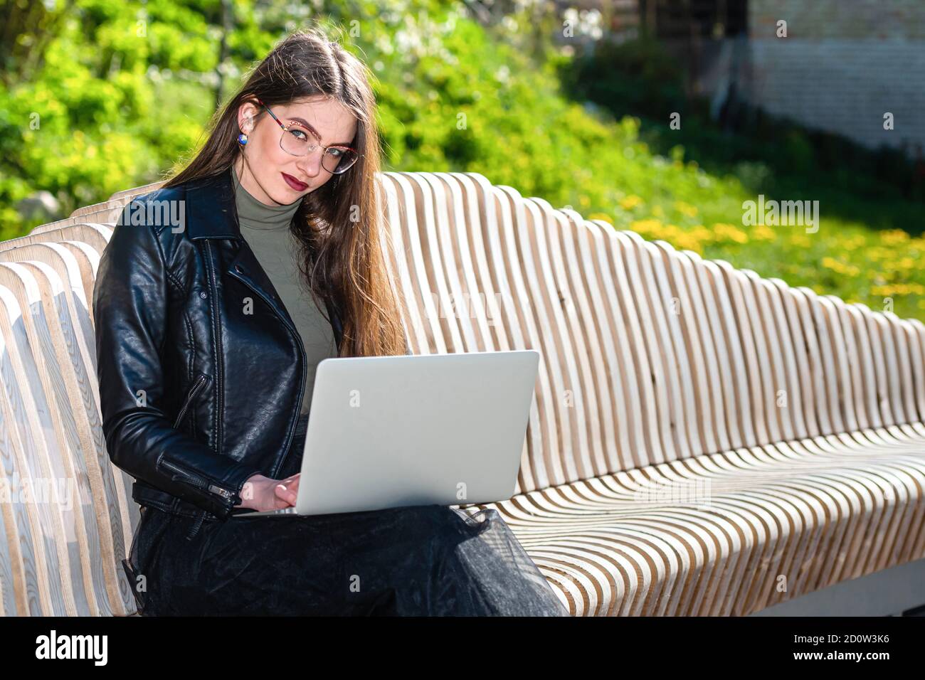 mujer joven en gafas sentada en un banco en el parque con ordenador portátil y mirando la cámara en un día soleado y ventoso de primavera, trabajando en cualquier lugar aw Foto de stock