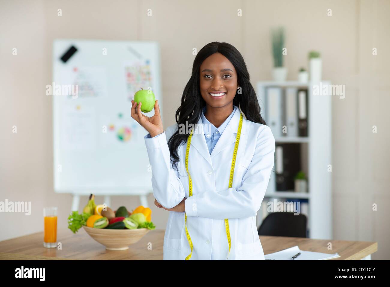 Mujer negra sonriente asesora de nutrición con manzana y cinta métrica mirando la cámara en la clínica Foto de stock