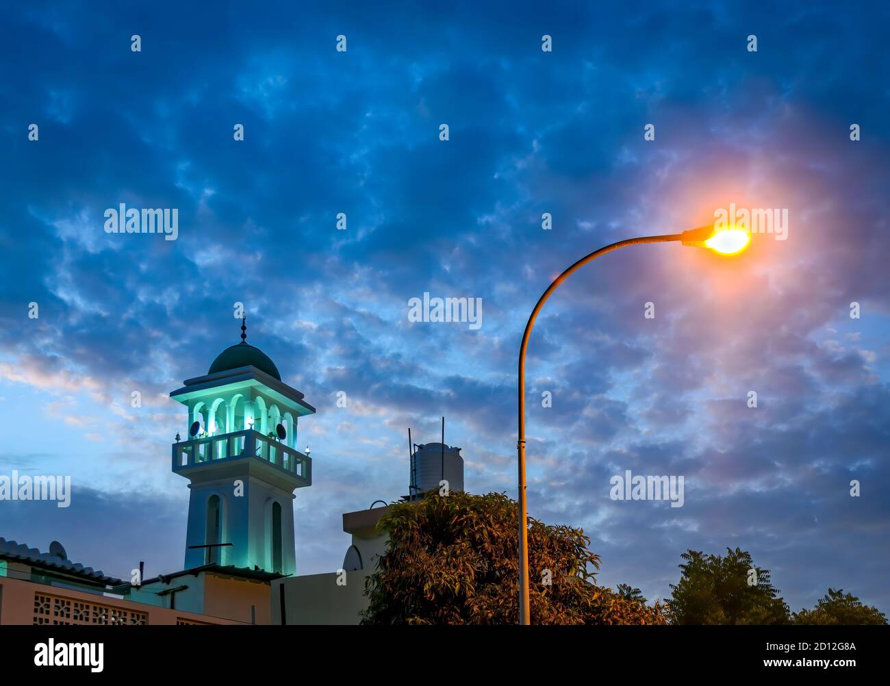 Torre de la Mezquita iluminada en verde contra un cielo azul por la noche alongwith un farol amarillo brillante. Desde Muscat, Omán. Foto de stock