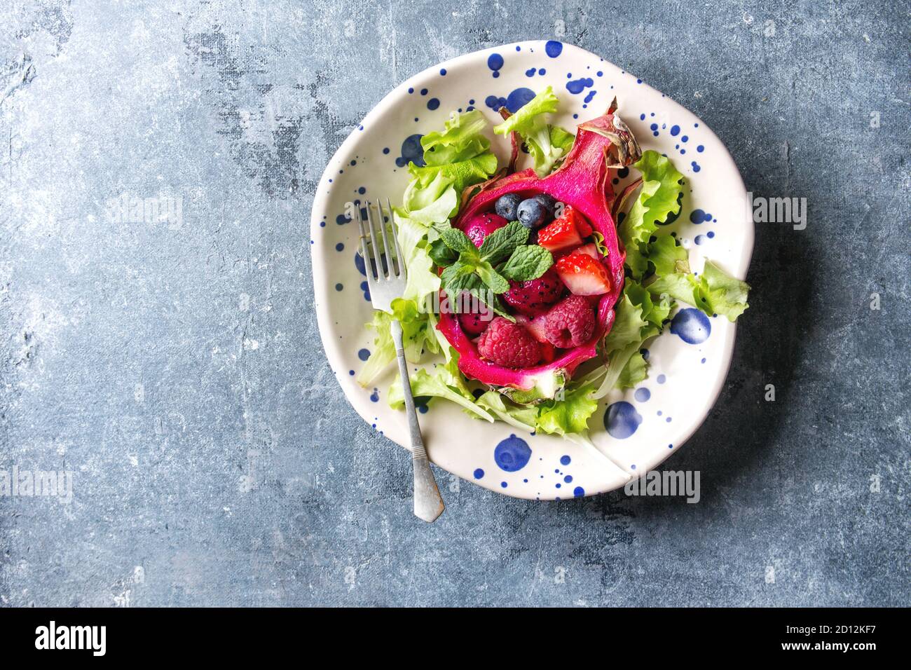 Ensalada de frutas veganas con bayas, ensalada verde y menta servida en fruta de dragón rosa en placa de cerámica manchada sobre fondo de textura azul. Vista superior, copiar Foto de stock