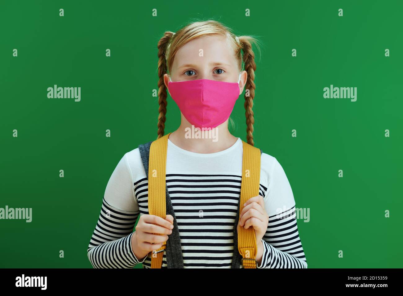 La vida durante la pandemia de covid-19. Retrato de la chica moderna de la escuela en blusa blanca con mochila amarilla y máscara rosa aisladas en el fondo verde pizarra Foto de stock