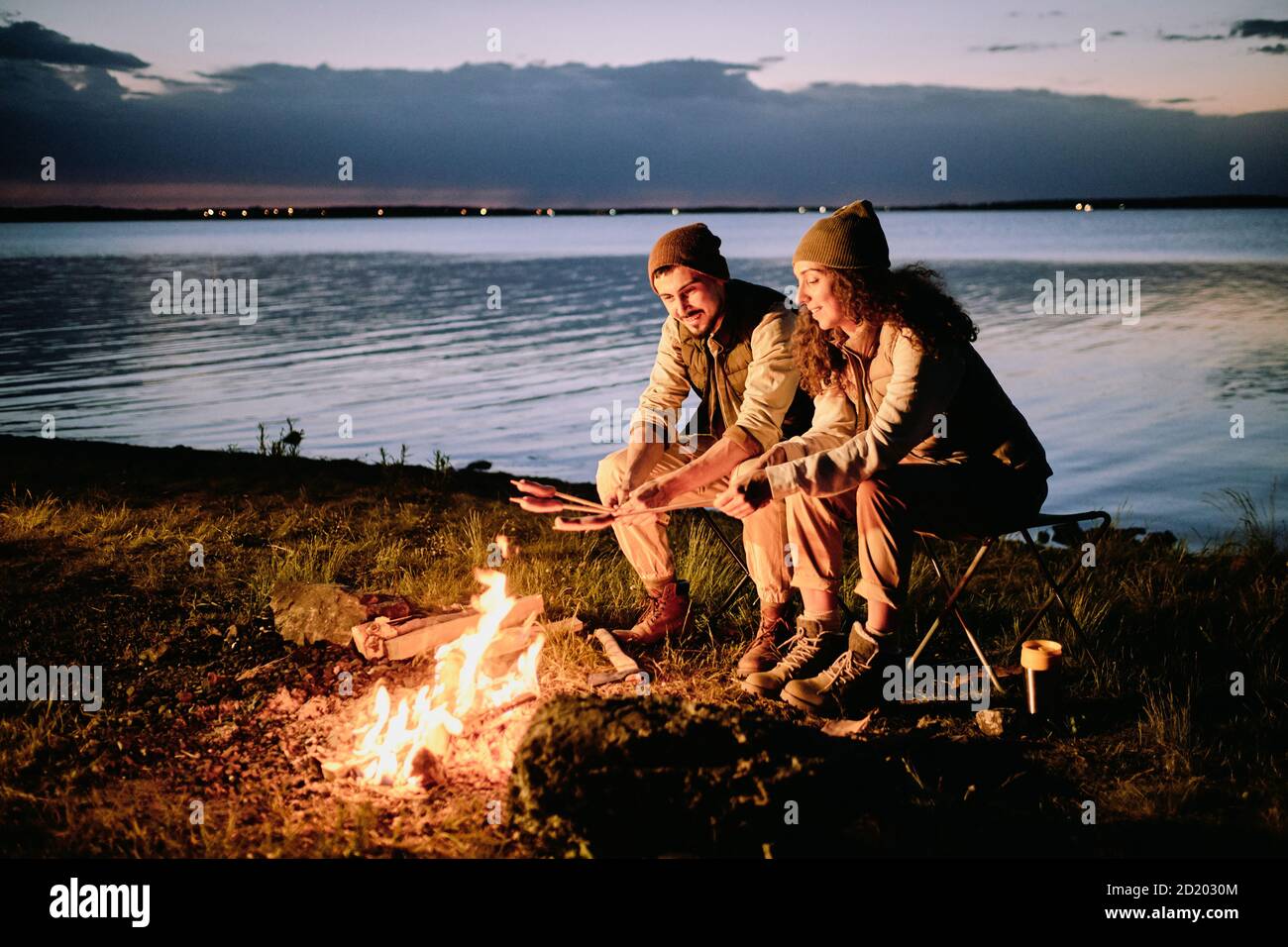 Una pareja joven positiva sentada en la orilla del lago y haciendo salchichas en el fuego durante el camping nocturno Foto de stock