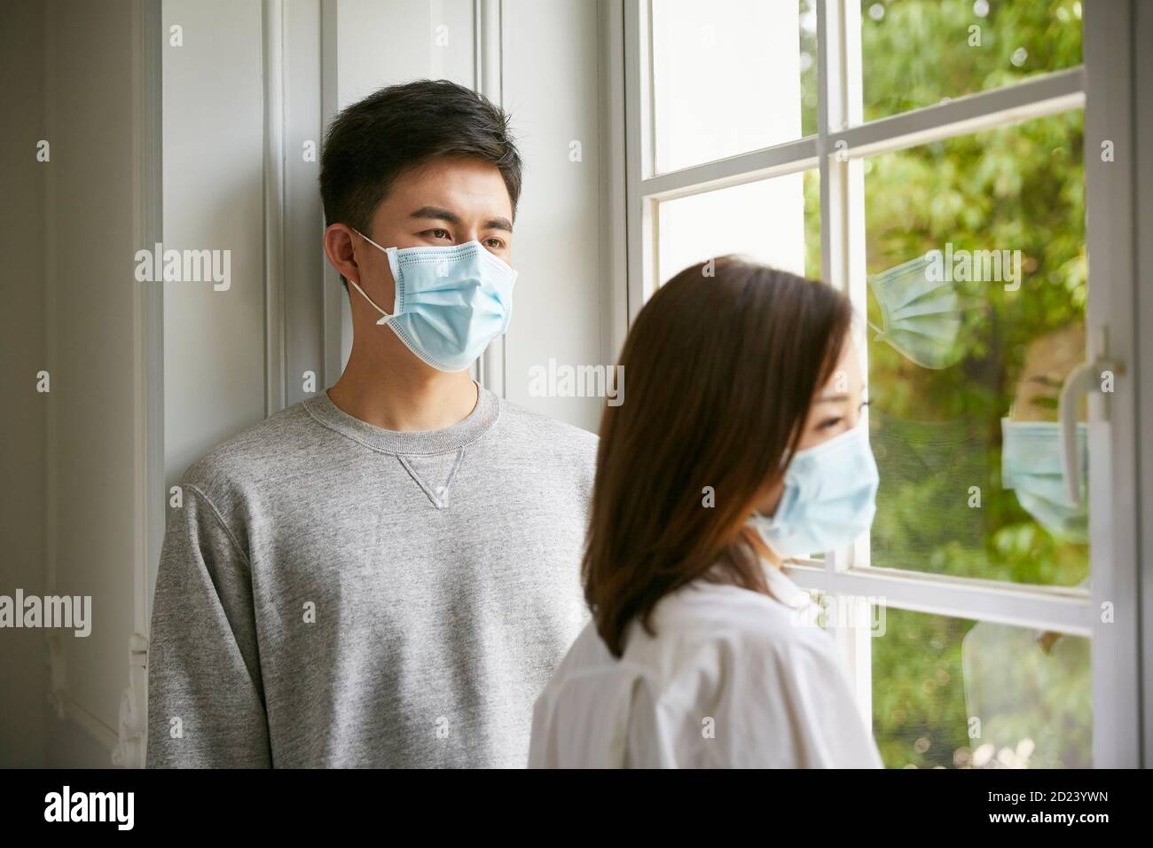 joven pareja asiática en aislamiento con máscara de pie junto a la ventana Foto de stock
