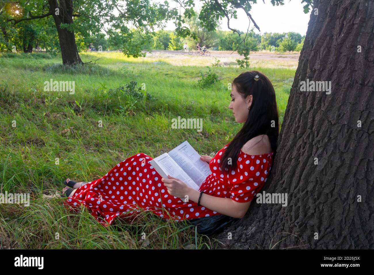 La joven está descansando en un día de verano en el parque. Una morena con un libro sobre la hierba cerca de un árbol se sienta con su espalda al tronco. Humor veraniego Foto de stock