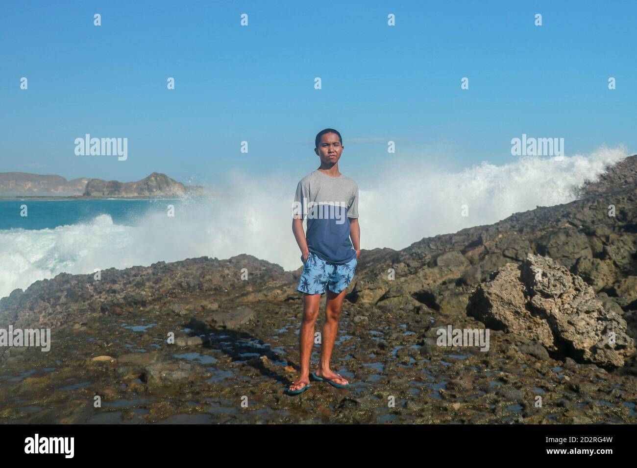 Olas golpeando rocas redondas y salpicando. Un joven está de pie en una orilla rocosa y las olas chocan contra un acantilado Foto de stock