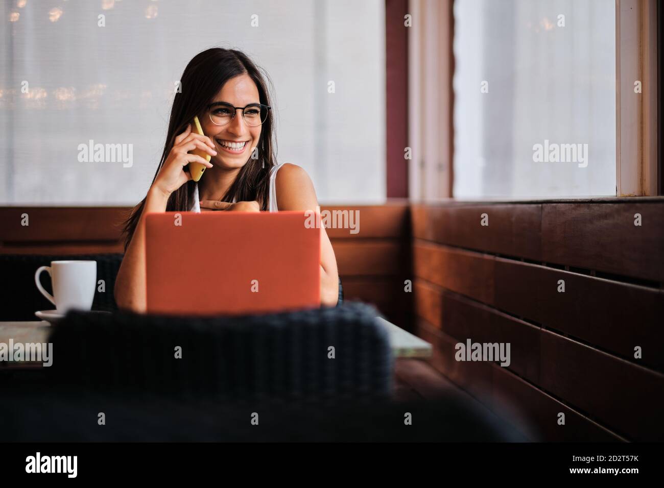 Una empresaria encantada trabajando remotamente en el café mientras discutía negocios problemas con el smartphone y sentarse en la mesa con el ordenador portátil Foto de stock