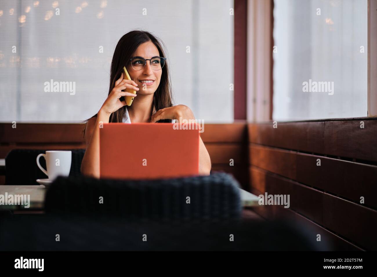 Una empresaria encantada trabajando remotamente en el café mientras discutía negocios problemas con el smartphone y sentarse en la mesa con el ordenador portátil Foto de stock