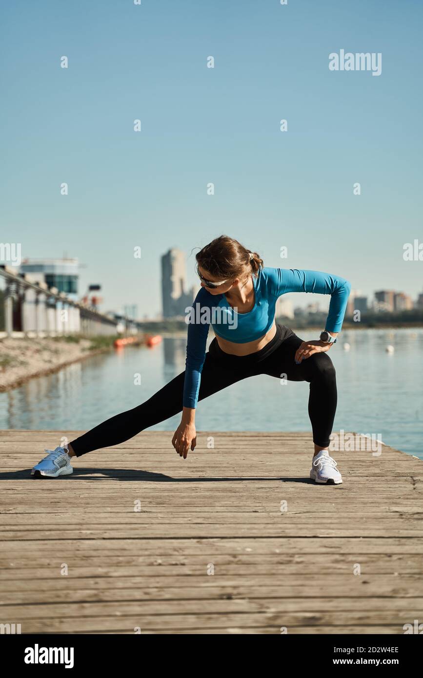 Mujer activa delgada en conjunto deportivo de pie sobre la alfombra y.. hacer  ejercicio de estiramiento para las piernas mientras entrena en el muelle de  madera cerca del río Fotografía de stock -