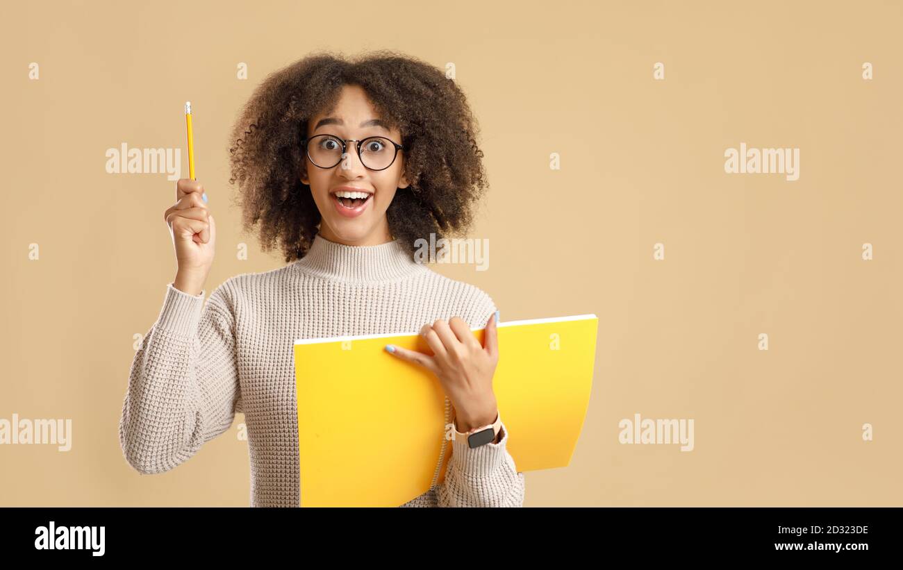 Gran idea para proyectos de educación o formación. Una mujer afro-americana sorpresa con gafas, con boca abierta y bloc de notas, levanta el lápiz Foto de stock