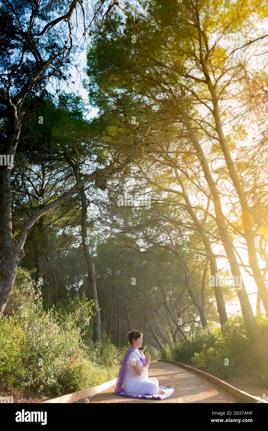 La mujer meditando sentada en el loto pose con sus manos delante de su pecho, en anjali mudra, en un camino del bosque al atardecer Foto de stock