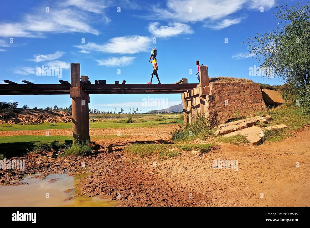 Ambalavao, Madagascar - 27 de abril de 2019: Desconocido Malagasy niño llevando la bolsa en su cabeza sobre un simple puente, otro sonriendo detrás de él, típico Mada Foto de stock