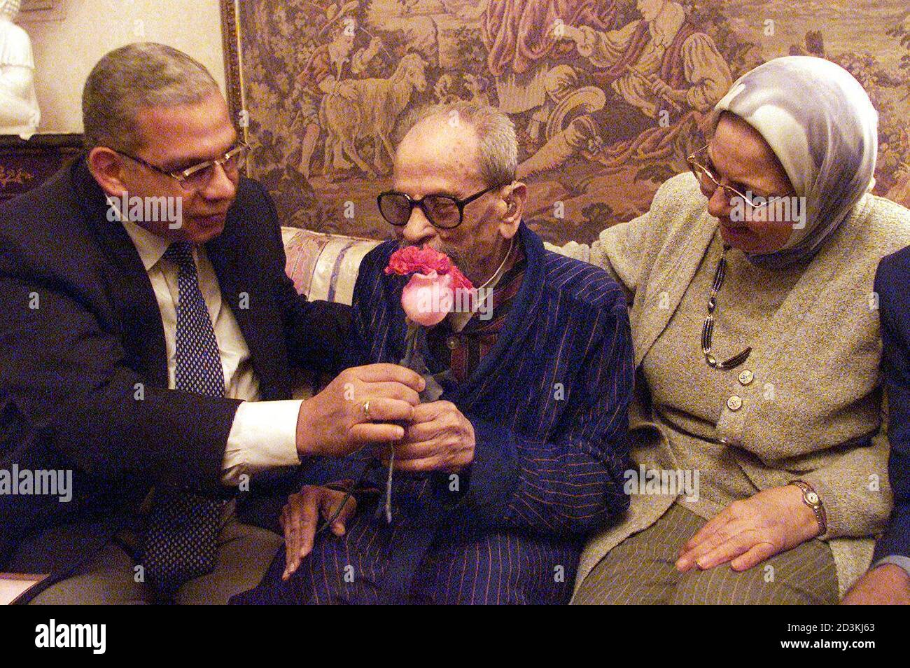Naguib Mahfouz (C), premio Nobel de Literatura de Egipto, recibe una flor del editor Mohammed Rashad mientras Salwa al-aanani, autor de su última biografía, mira un día antes de su 90 cumpleaños en su casa en el Cairo el 10 de diciembre de 2001. Mahfouz se convirtió en el primer escritor en árabe en ganar el premio Nobel en 1988. En 1994 fue apuñalado por un militante musulmán en un intento fallido de asesinato, después de que los militantes lo declararan infiel por su imagen de Dios en la novela "los hijos de Gebelawi". REUTERS/Aladin Abdel Naby AN Foto de stock