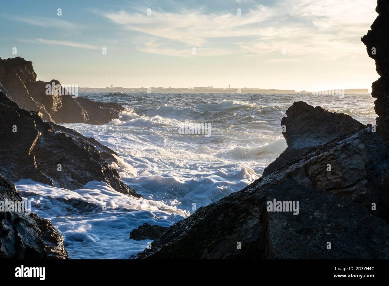 Mar espumoso con olas y rocas en primer plano al atardecer con luz cálida y Peniche península en la distancia, Baleal Portugal Foto de stock