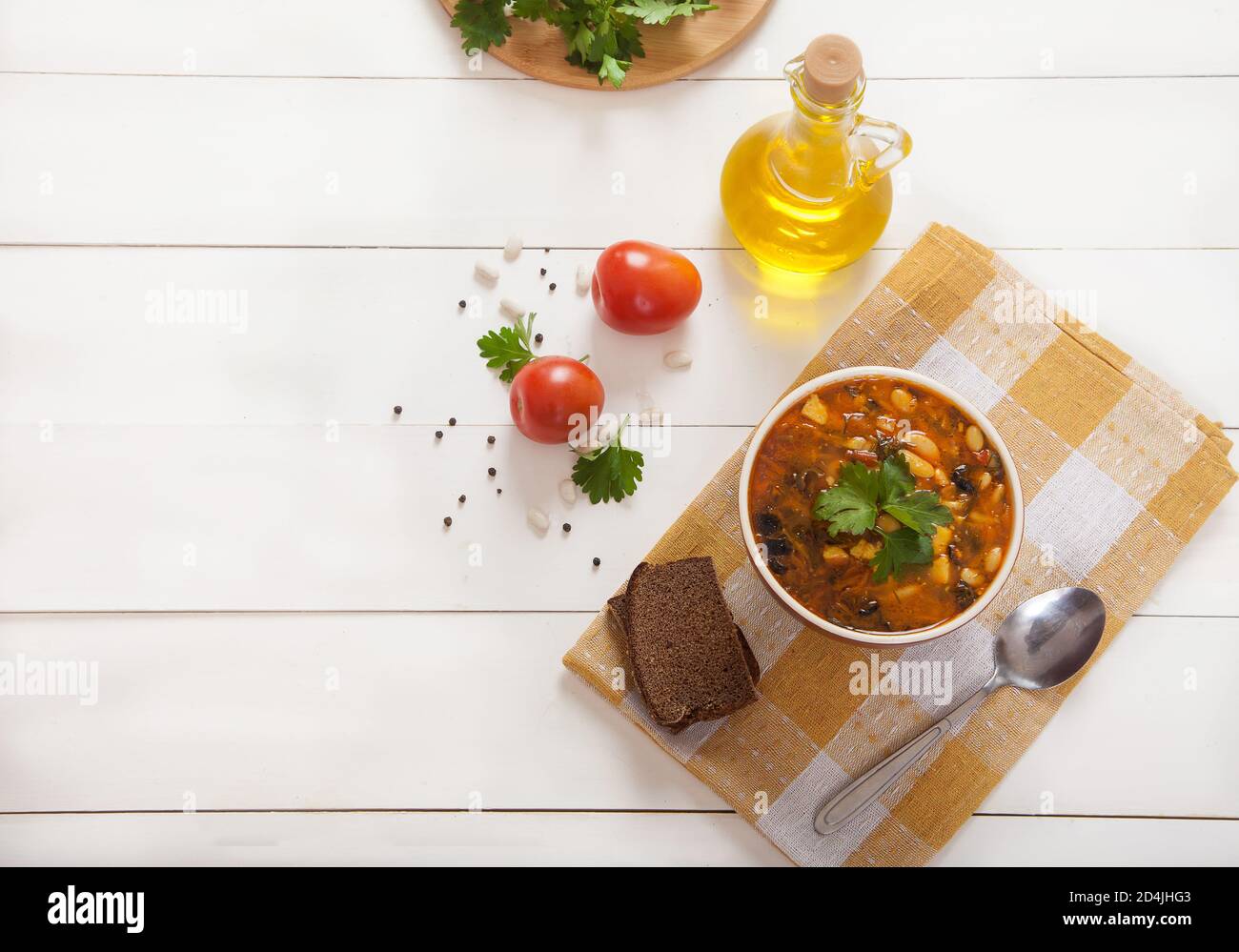 Sopa vegetariana con frijoles y aceitunas y tomates en una taza de cerámica, aceite de oliva en una servilleta de lino amarillo sobre un fondo de madera blanco. Copiar perchas. Foto de stock
