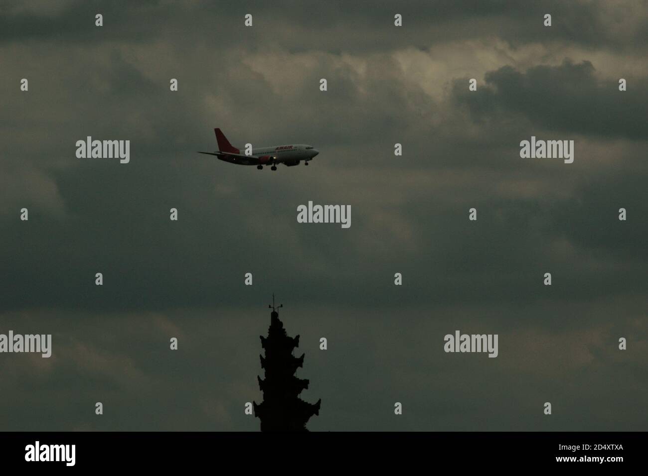 Un avión indonesio AWAIR volando sobre el Aeropuerto Internacional Soekarno-Hatta de Yakarta, con la silueta de una torre de puerta de bienvenida construida en estilo arquitectónico balinés en el lado de la carretera de peaje del aeropuerto se ve en parte. Foto de stock