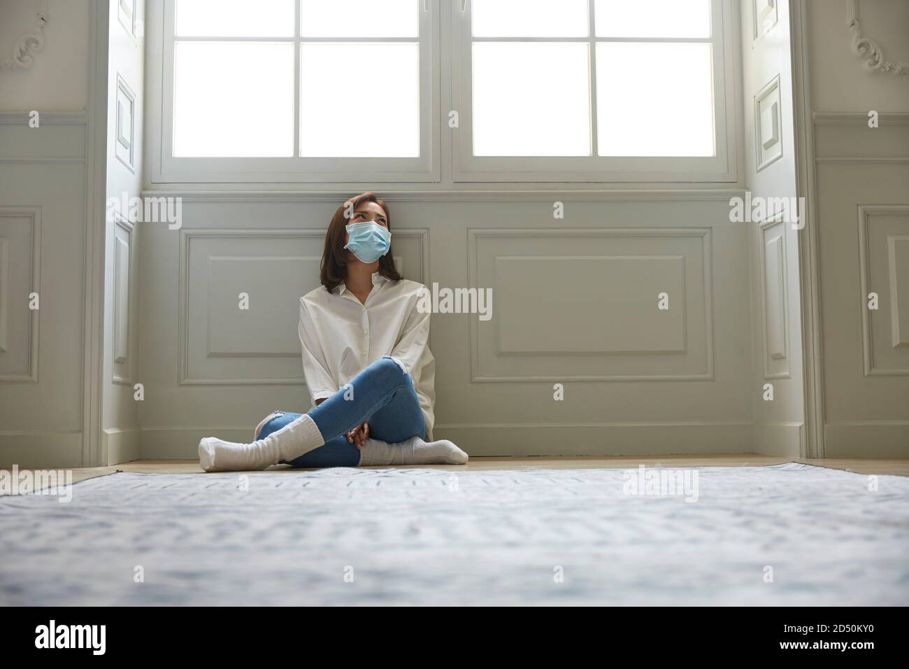 mujer asiática joven en cuarentena en casa usando máscara facial sentado en las piernas del piso se cruzó con un aspecto triste y deprimido Foto de stock
